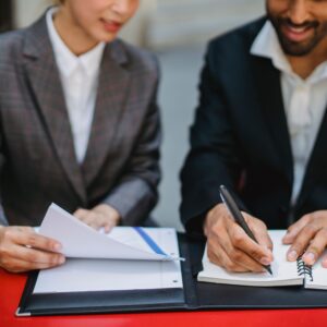Two business professionals in suits reviewing documents and writing notes at a table.